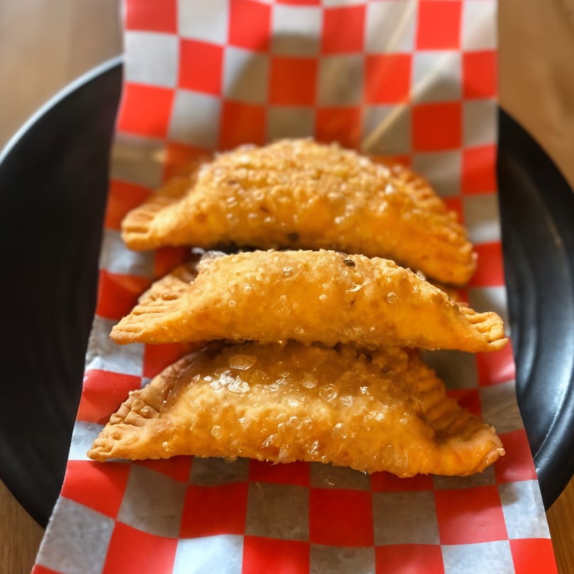Three golden-brown Peruvian empanadas, sprinkled with salt, rest on red and white checkered paper.