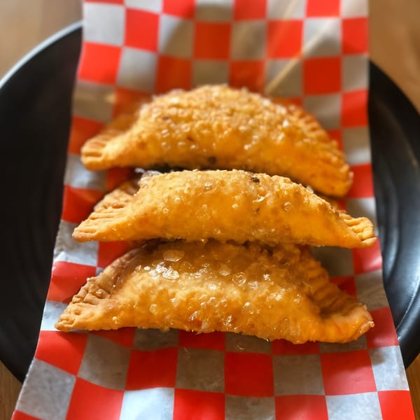 Three golden-brown Peruvian empanadas, sprinkled with sugar, stacked on red and white checkered paper.
