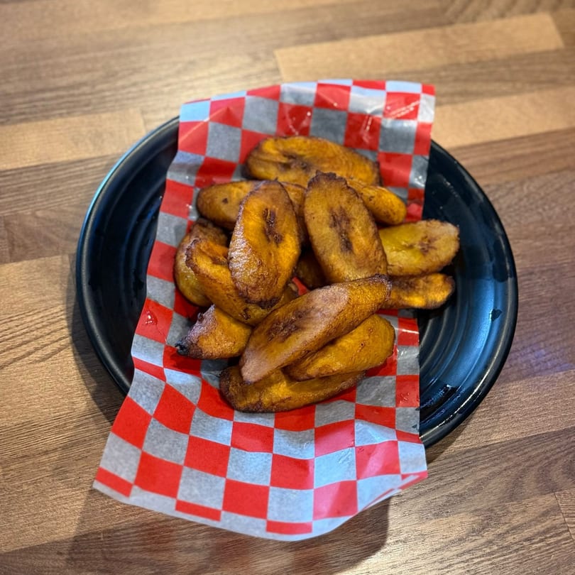 A black plate holds golden-brown fried plantains on red and white checkered paper.
