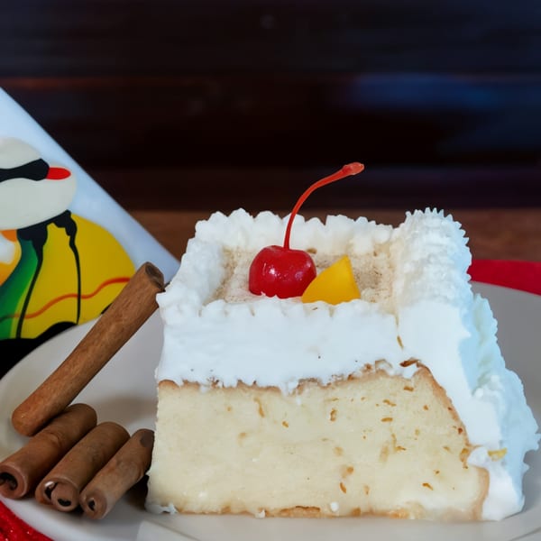 A slice of Peruvian tres leches cake with whipped cream, a cherry, and cinnamon sticks.